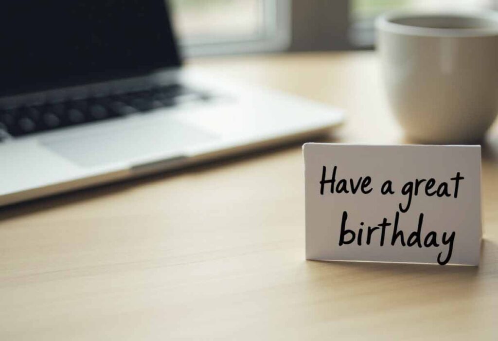 Office desk with card reading “Have a great birthday” beside coffee cup and laptop.