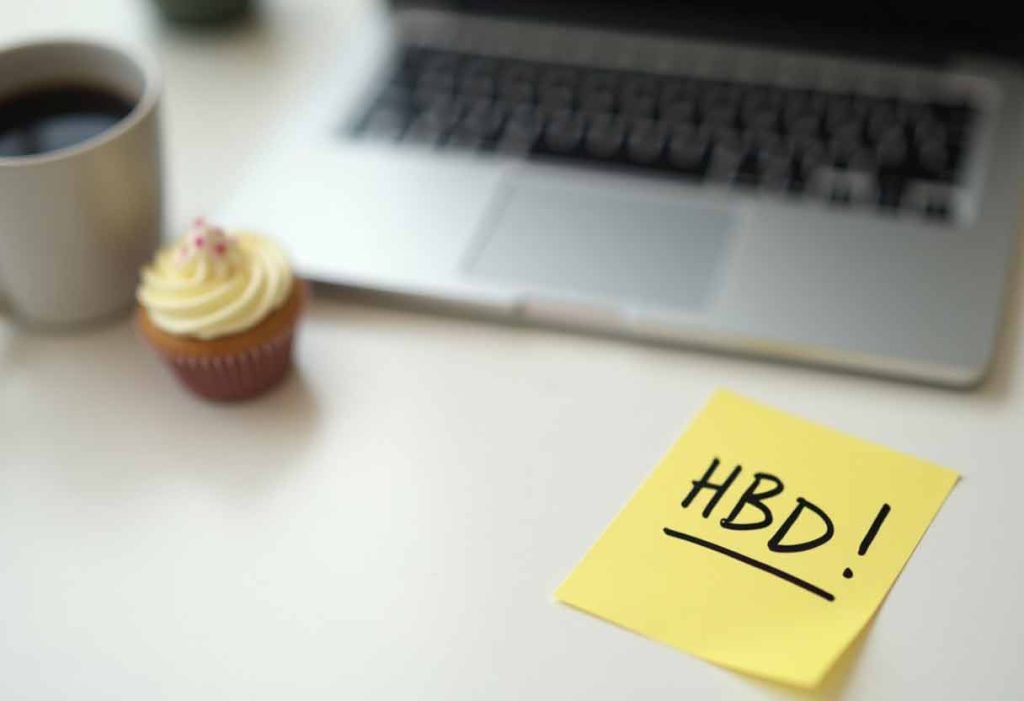 Work desk with closed laptop, coffee mug, cupcake, and a sticky note that says “HBD!”