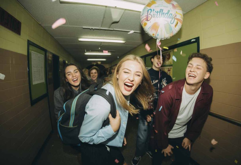 High-school hallway with students laughing mid-joke as confetti falls and a birthday balloon bounces.