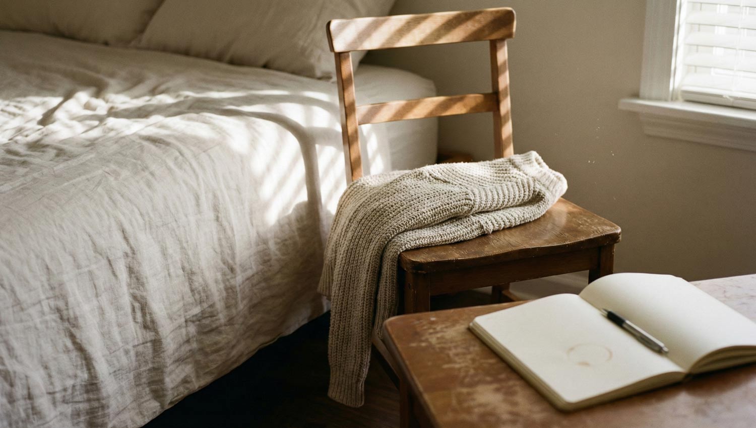 A quiet morning interior scene showing subtle routine: a neatly made bed, sunlight falling across a chair with a folded sweater, a notebook and pen on a table. No people visible, but human presence implied. Soft, warm light, muted colors, realistic textures. Feels like slow progress, gentle stability, and private rebuilding. Editorial photography style.