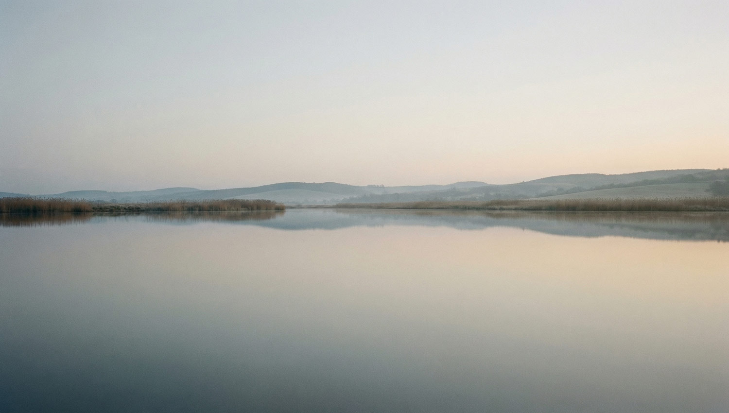 A still body of water at dawn with no people present, subtle reflections, pale sky. Natural landscape, realistic photography style, calm and unobserved feeling, no dramatic colors or contrast.