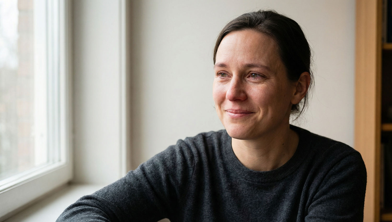 Realistic portrait of a person smiling while their eyes show quiet emotion. Soft daylight from a window, neutral background, minimal styling. Expression balances humor and sincerity. Natural imperfections, subtle shadows, emotional realism, modern editorial photography style.