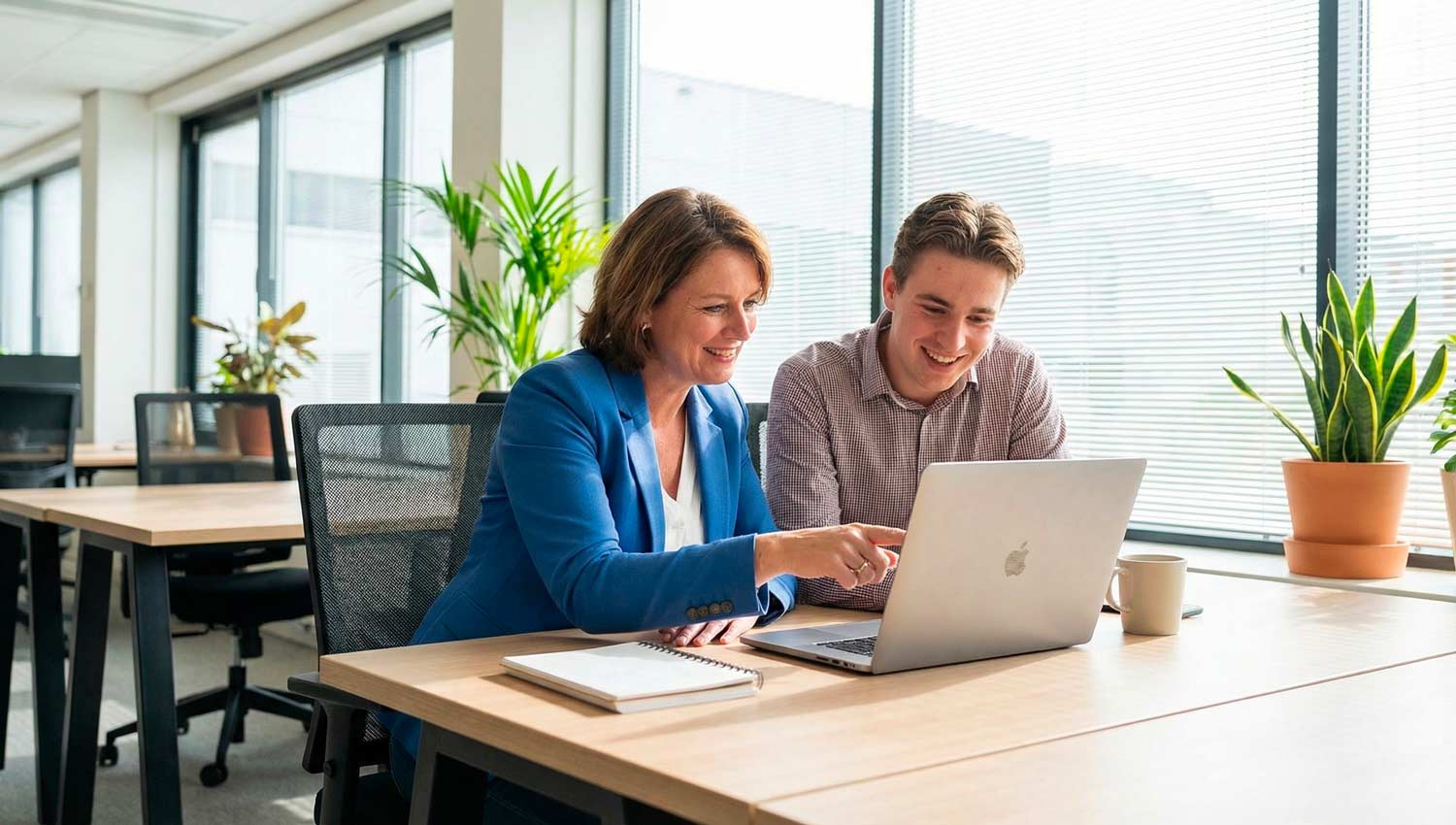 A modern office setting where a mentor and mentee sit side by side reviewing work on a laptop. The mentor has a calm, guiding presence, slightly leaning forward while explaining something, while the mentee listens attentively with respect. Natural daylight enters through large windows, creating a professional yet warm atmosphere. Minimalist desk setup, soft shadows, realistic human expressions, cinematic lighting, no text, no logos.