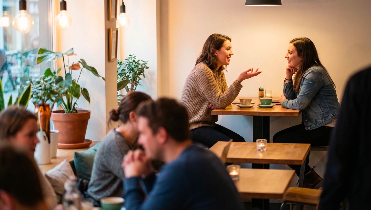 Two close friends sitting together in a cozy café, laughing and talking deeply over coffee. One friend gestures while sharing something meaningful, the other listens with genuine interest. Warm ambient lighting, soft reflections, modern café interior with plants and wooden textures. The scene captures authentic connection, growth, and friendship energy. Ultra-realistic, cinematic warmth, no text, no logos.