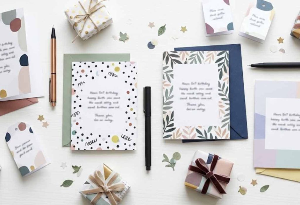 A photorealistic flat-lay of several small birthday cards, gift tags, colorful envelopes, pens, and miniature wrapped presents on a clean white tabletop with subtle confetti.